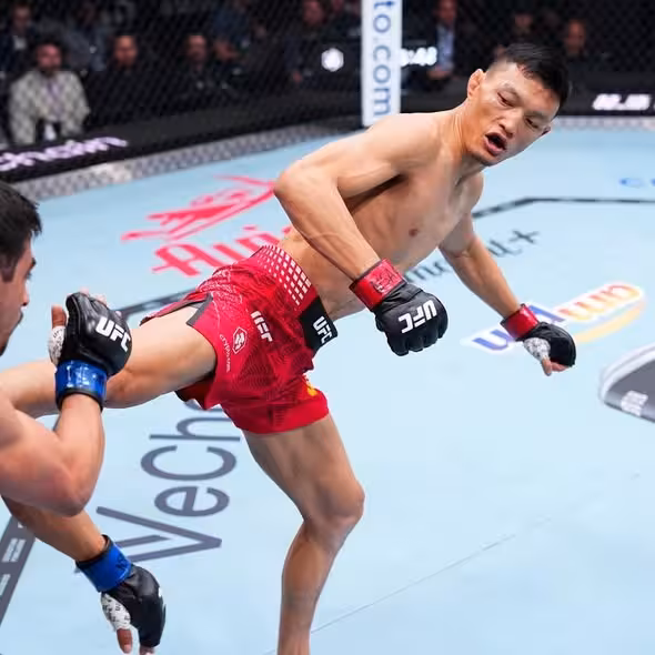 UFC fighter Su Mudaerji executing a dynamic high kick with his right leg against an opponent during a bout inside the octagon